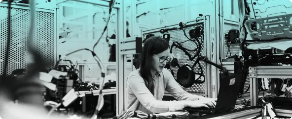 A person working at a laptop inside a workshop, surrounded by machinery and equipment racks