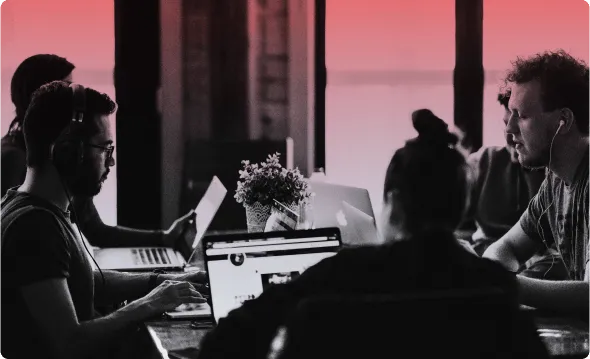 Several people working on laptops at a long shared table, headphones on